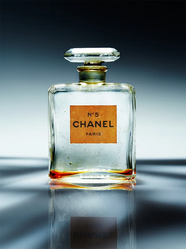 Fotografia concettuale di una bottiglia di profumo Chanel N°5 vintage con etichetta arancione, posta su una superficie riflettente con sfondo grigio sfumato.