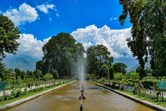Jardines Shalimar en Cachemira con una fuente central, árboles, caminos con flores y un cielo azul con nubes.