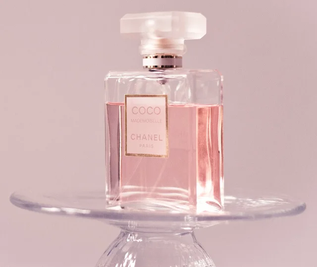 Botella de perfume Coco Mademoiselle de CHANEL con líquido rosado, posada sobre un soporte de vidrio transparente, con fondo rosado.