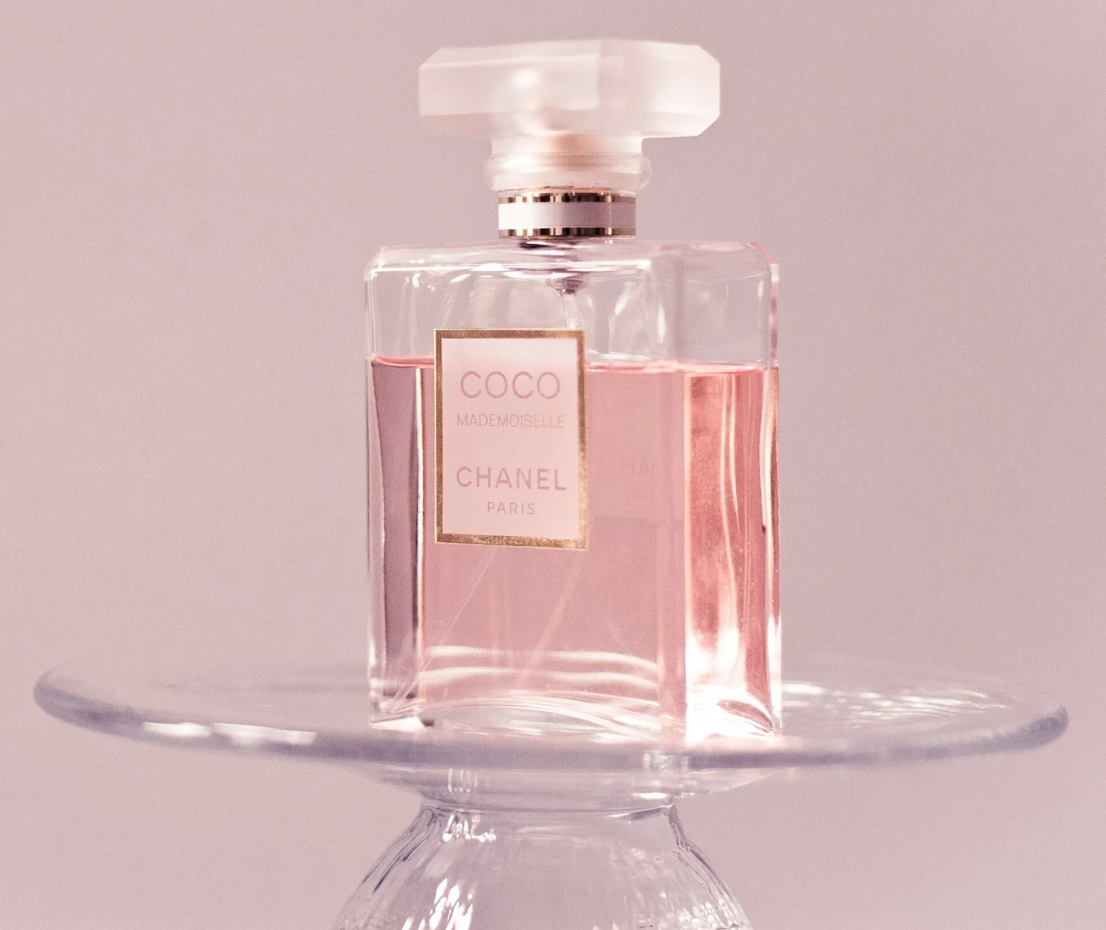 Botella de perfume Coco Mademoiselle de CHANEL con líquido rosado, posada sobre un soporte de vidrio transparente, con fondo rosado.