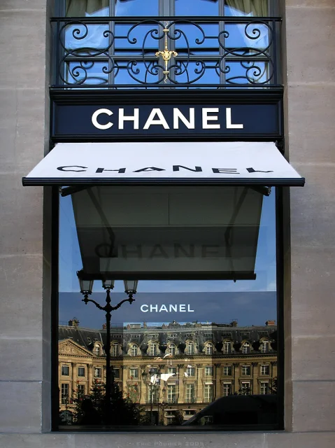 Fachada de la boutique Chanel Joaillerie en Place Vendôme, París, con el logo de Chanel visible y reflejo del edificio frente a la ventana.