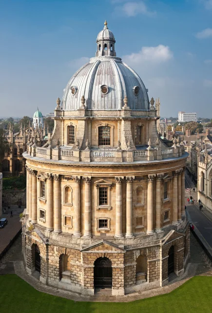 Fotografía del edificio Radcliffe Camera en Oxford, Inglaterra, con su icónica cúpula y arquitectura clásica sobre un césped verde bajo un cielo azul claro.