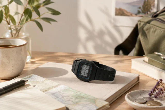 Imagen conceptual de un reloj digital CASIO F-91W-1 colocado sobre un cuaderno en una mesa de madera, con una taza, un bolígrafo, un mapa, monedas y plantas de fondo.